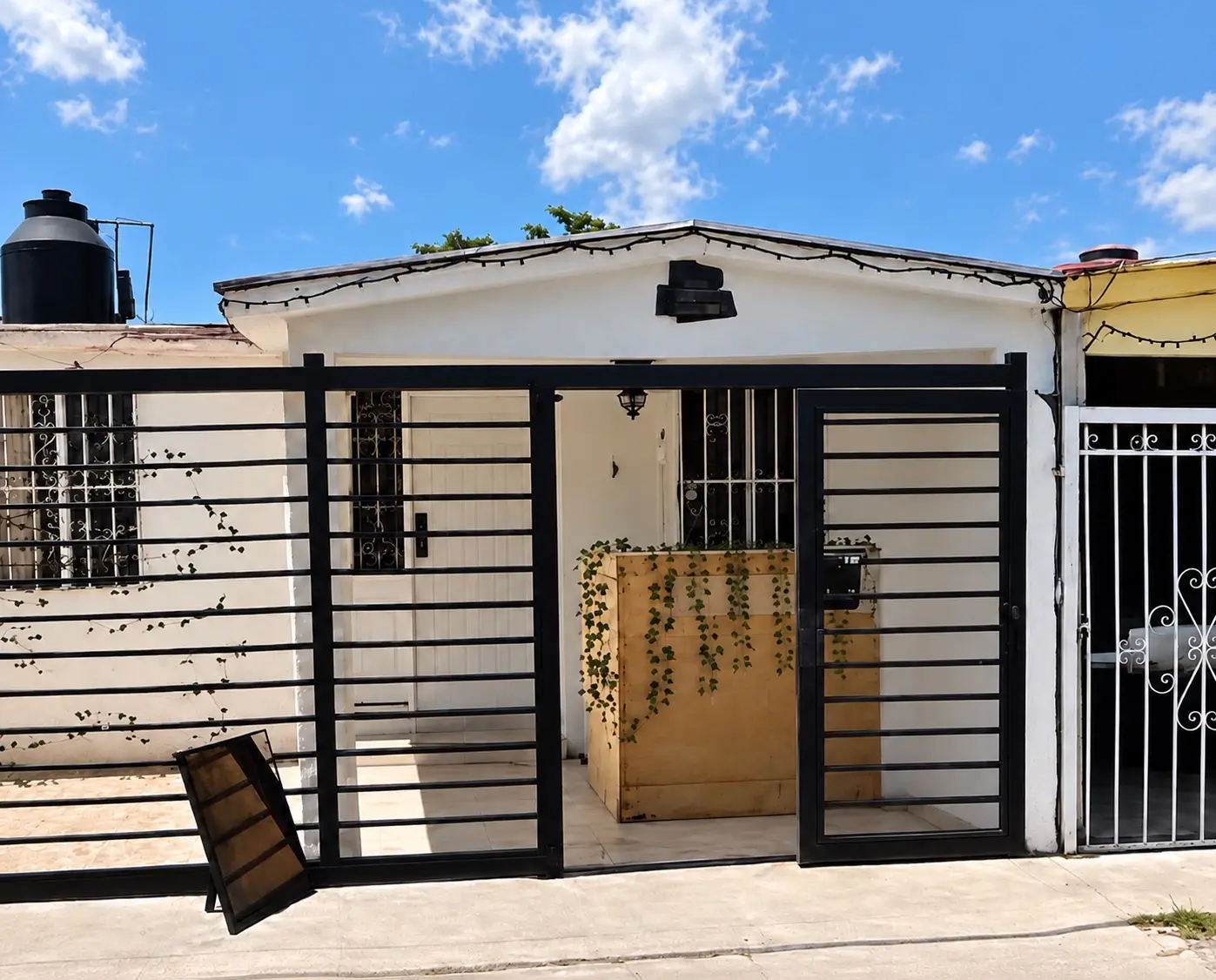 La cocherita donde empezó andy & dany: una casa blanca con reja negra, foquitos en el techo y una barra de madera con enredadera en la cochera
